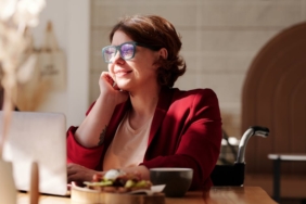Cheerful woman in a red blazer working remotely on a laptop at a café, enjoying the sunlight.
