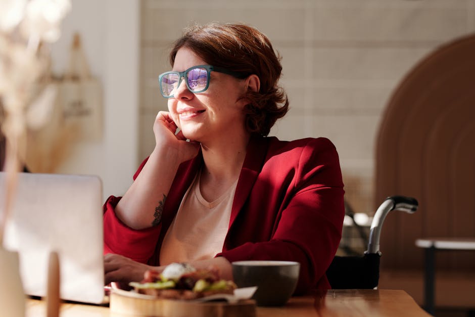 Cheerful woman in a red blazer working remotely on a laptop at a café, enjoying the sunlight.