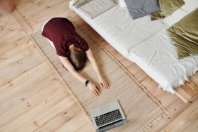Woman in yoga pose on mat, indoors with laptop, showing mindfulness and home fitness.