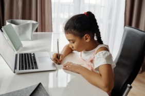 A girl focusing on homework, writing at a desk next to a laptop.