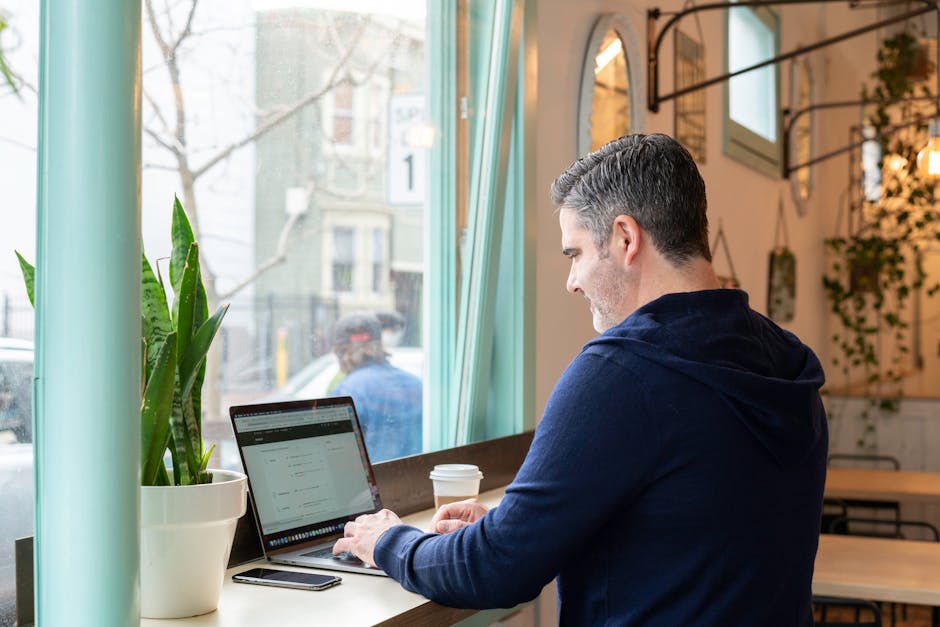 Man working remotely on laptop in a cozy urban cafe, enjoying a coffee with a view outside.