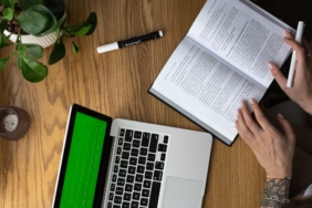 From above of crop anonymous tattooed female student reading book while doing homework assignment sitting at wooden table with laptop and potted plant