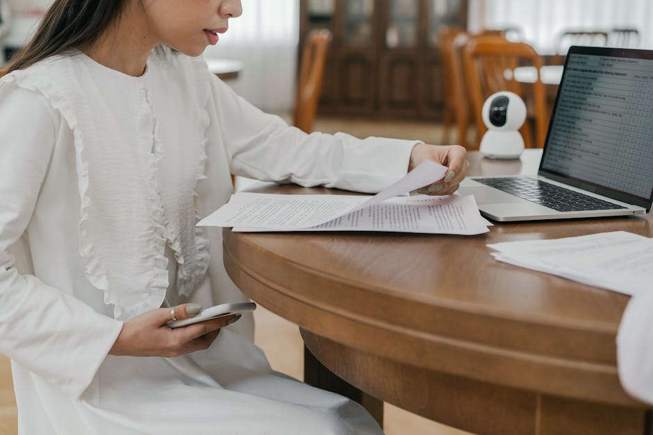 Woman in white dress studying at a wooden table with a laptop and papers indoors.