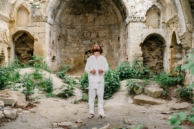 A man in white clothing stands in historic stone ruins with lush greenery.